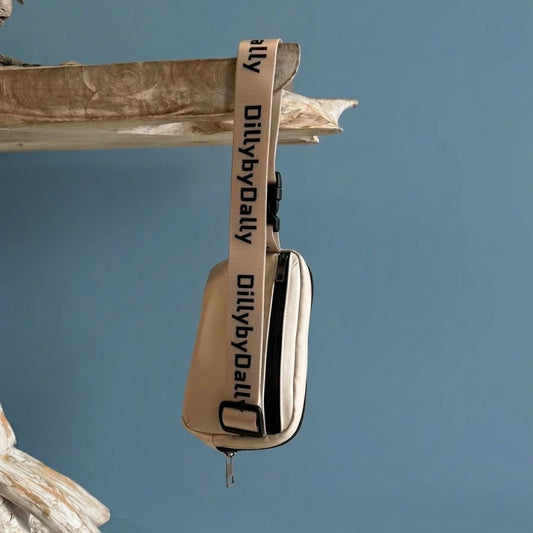 A beige and black crossbody bag with a wide beige strap printed with 'DillybyDailyy' in black, hanging from a rustic wooden branch against a solid blue background.