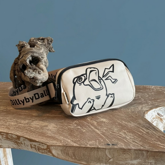 A cream-colored crossbody bag with a black line-art illustration of an overstimulated Dilly the platypus and a black-and-white patterned strap, styled on a rustic wooden table next to a piece of ornate wood against a solid blue-gray background.