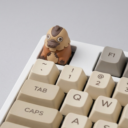 A close-up of a beige mechanical keyboard with a small cartoon Dilly the platypus keycap inserted into the esc slot, set against a neutral gray background.